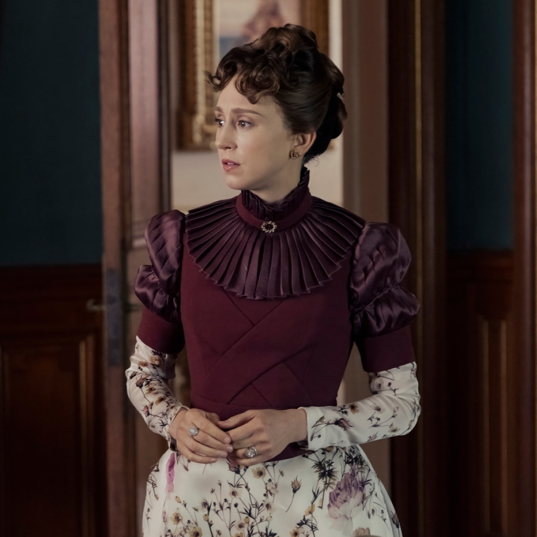 taissa farmiga as gladys russell wearing a maroon and floral dress standing in a parlor in the gilded age