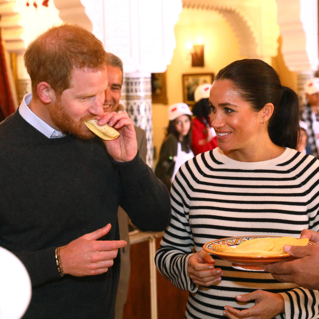 Prince Harry eats a sandwich while Meghan Markle, wearing a striped top, watches